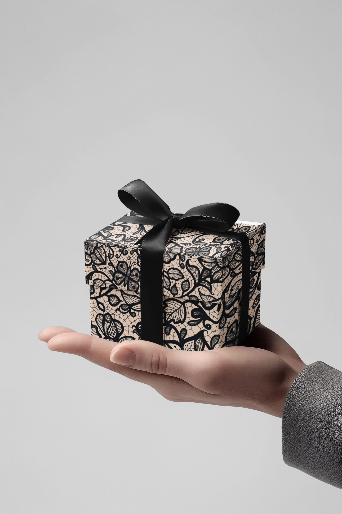 Hand holding a small gift box with black lace design and a black ribbon on a light gray background