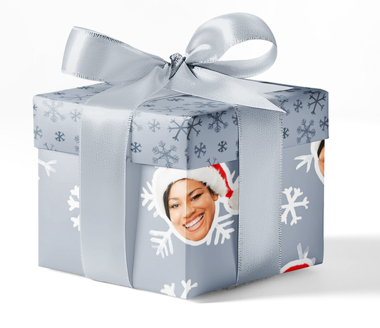 A gift box wrapped with custom wrapping paper featuring a snowflake pattern and a photo of a smiling person wearing a Santa hat.